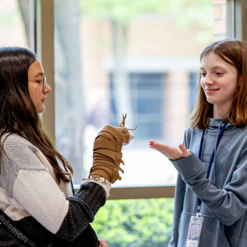 Two young students standing by a window, one holding a worm puppet while the other has her hand out to interact with it.